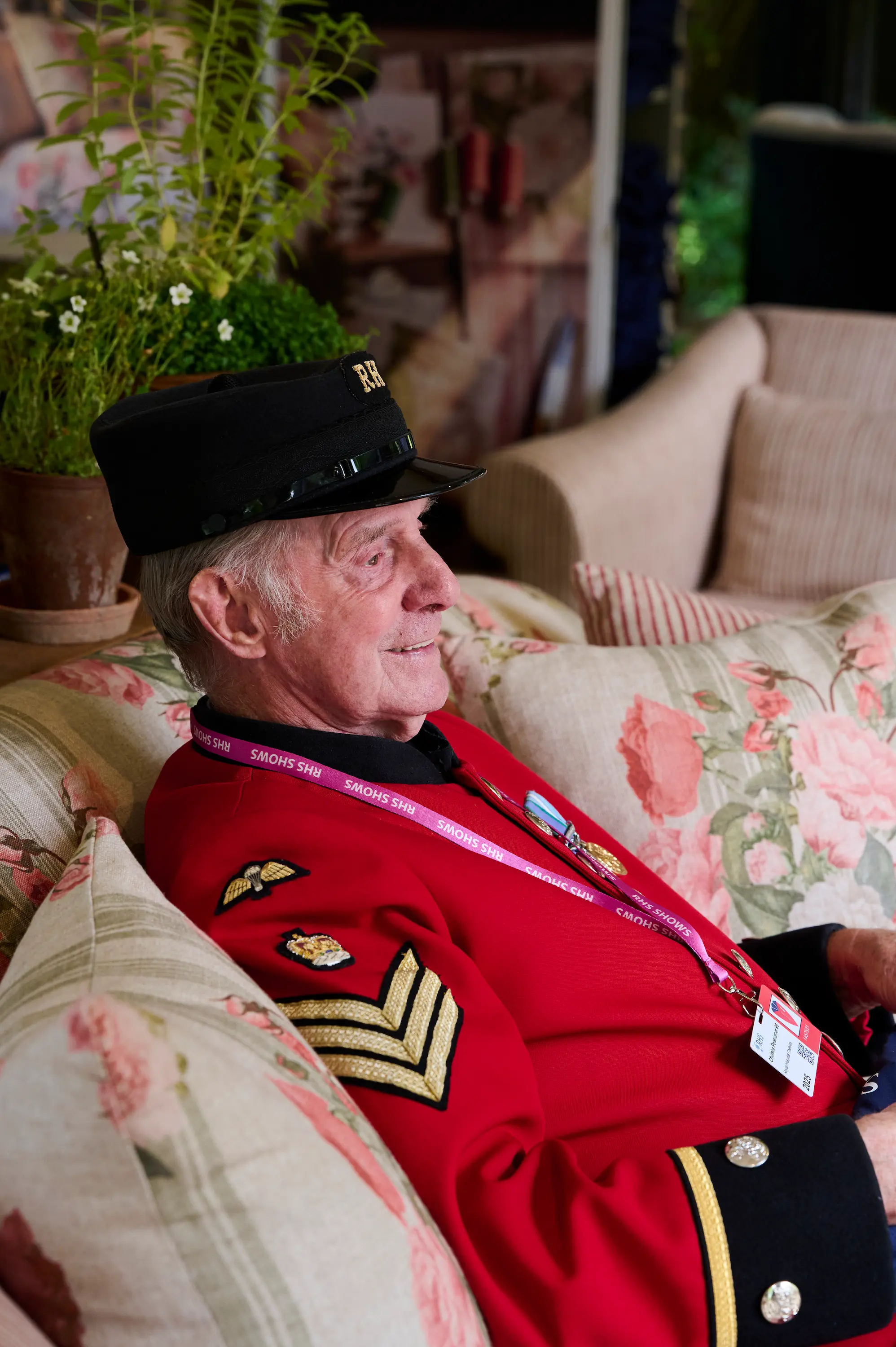 An elderly man in a red uniform and black hat sits on a floral sofa. He wears a lanyard with badges and medals on his uniform, and is surrounded by plants and floral decor.